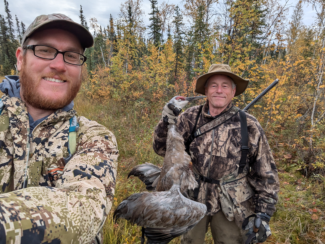 Two men dressed in camouflage in the backcountry of alaska. One man is smiling and holding a dead crane.